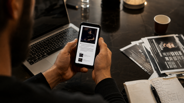 A person reviews a DJ's press kit on a smartphone, showing a photo of the DJ behind the decks, while a laptop and printed press photos sit on the desk nearby.