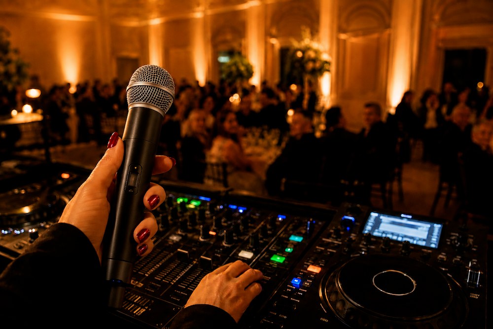 Mobile DJ mic technique in action as a DJ holds a wireless handheld mic above a lit mixer while guests dine in a ballroom