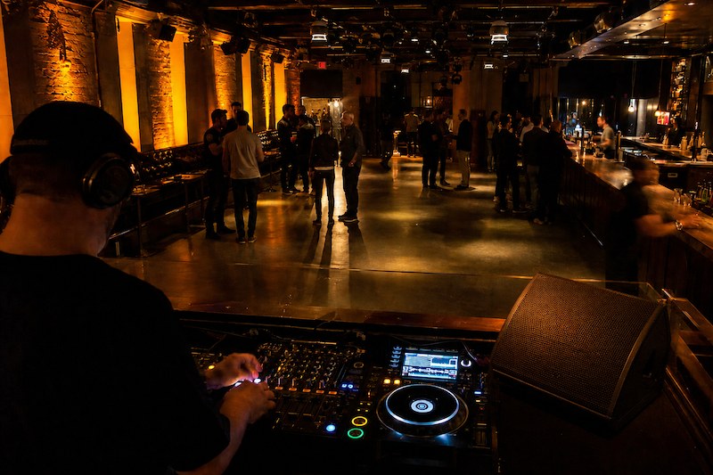 DJ playing an opening set from behind the booth as a club fills, warm amber lights and a nearly empty floor